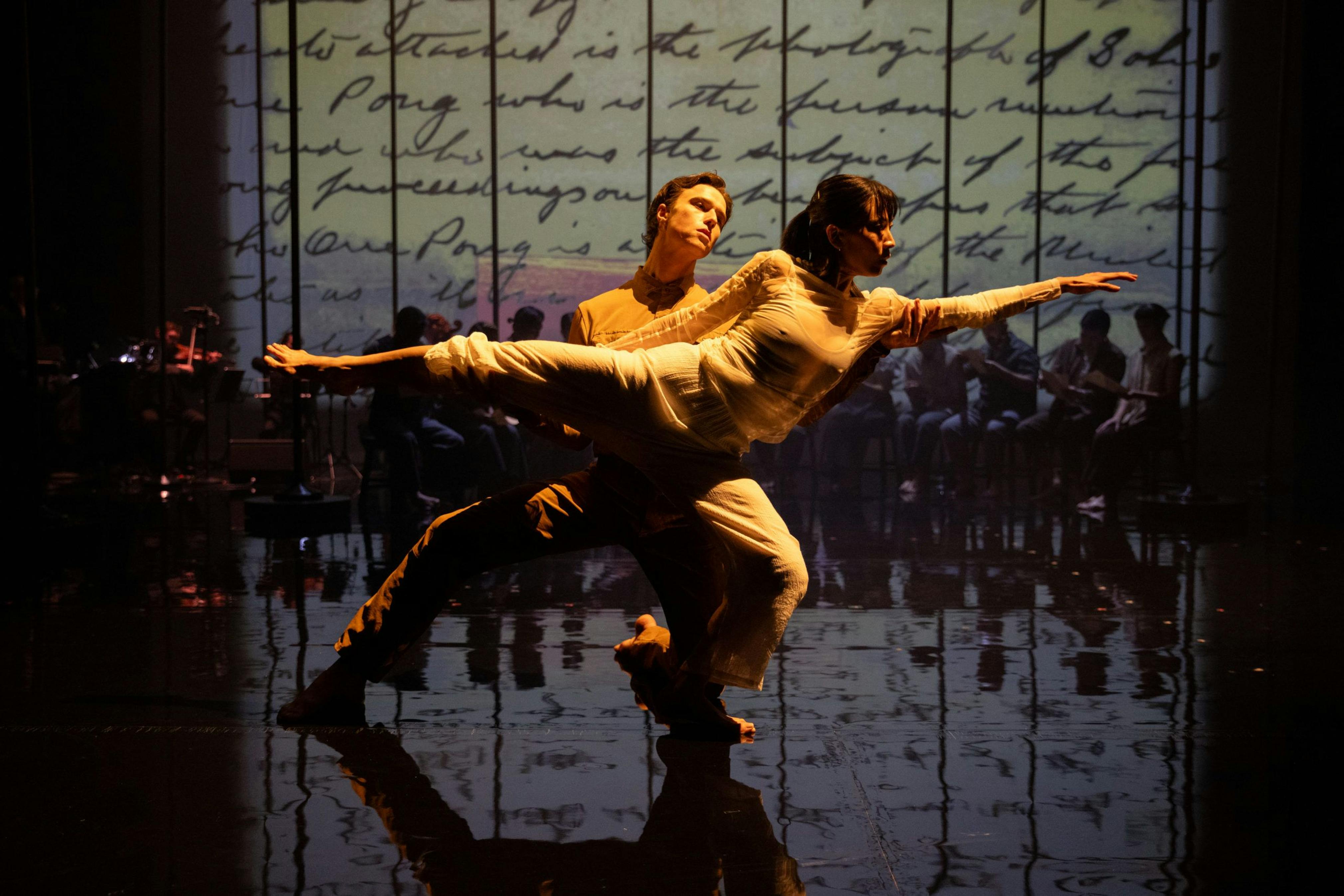 The image shows an old black and white photo hung on a black wall with barbwire. The photo displays several asian families touching shoulder to shoulder while militant men stare at them.